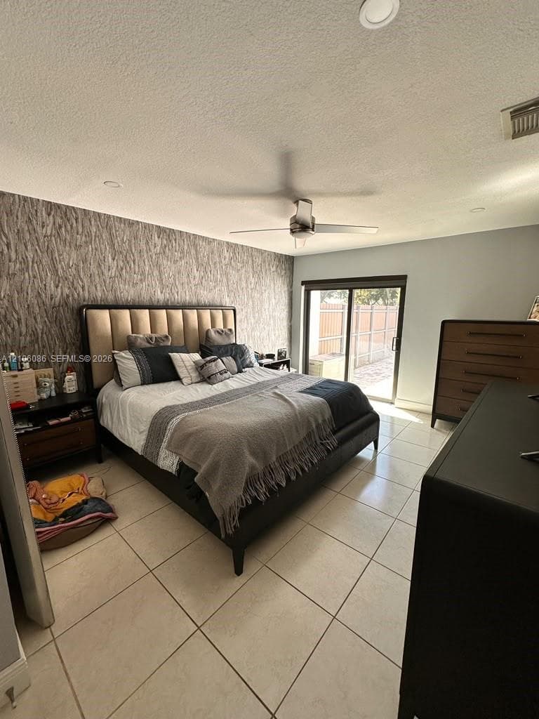 Room featuring textured accent wall, light-colored tile flooring, ceiling fan, and sliding glass door
