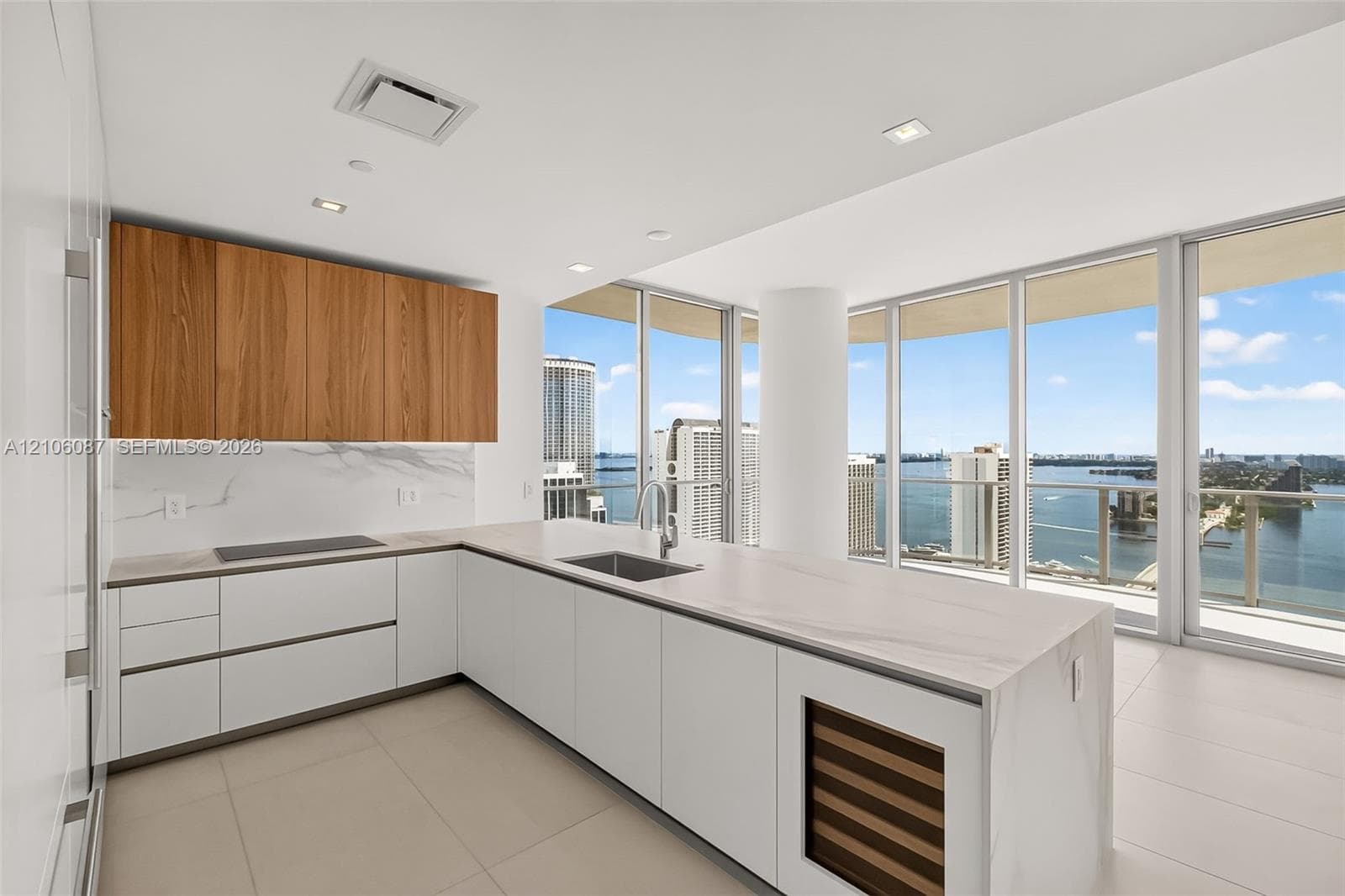 Kitchen featuring wood-finish upper cabinetry, white flat-panel lower cabinetry, light-toned stone-surface countertops, integrated wine refrigeration, and floor-to-ceiling glass windows