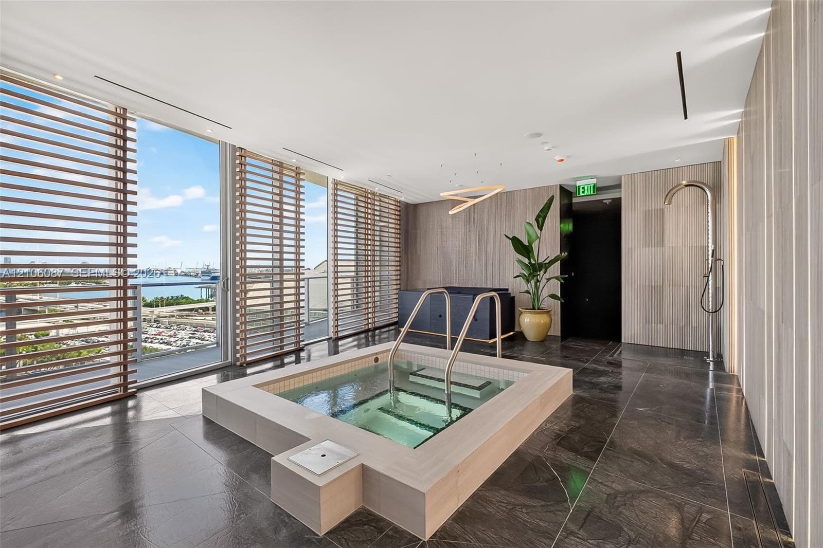 Recessed soaking tub featuring a stone-tile surround, floor-to-ceiling windows with wood-slat screening, large-format stone floor tiles, and a wall-mounted shower fixture