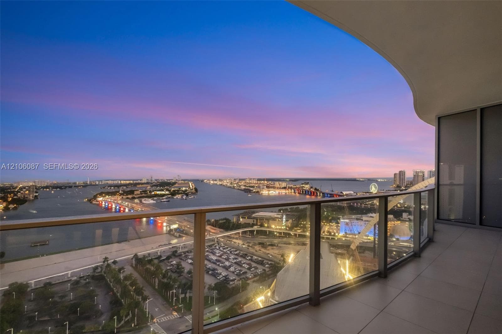 Curved balcony featuring large-format tile flooring and a glass-paneled railing system overlooking a coastal cityscape and harbor