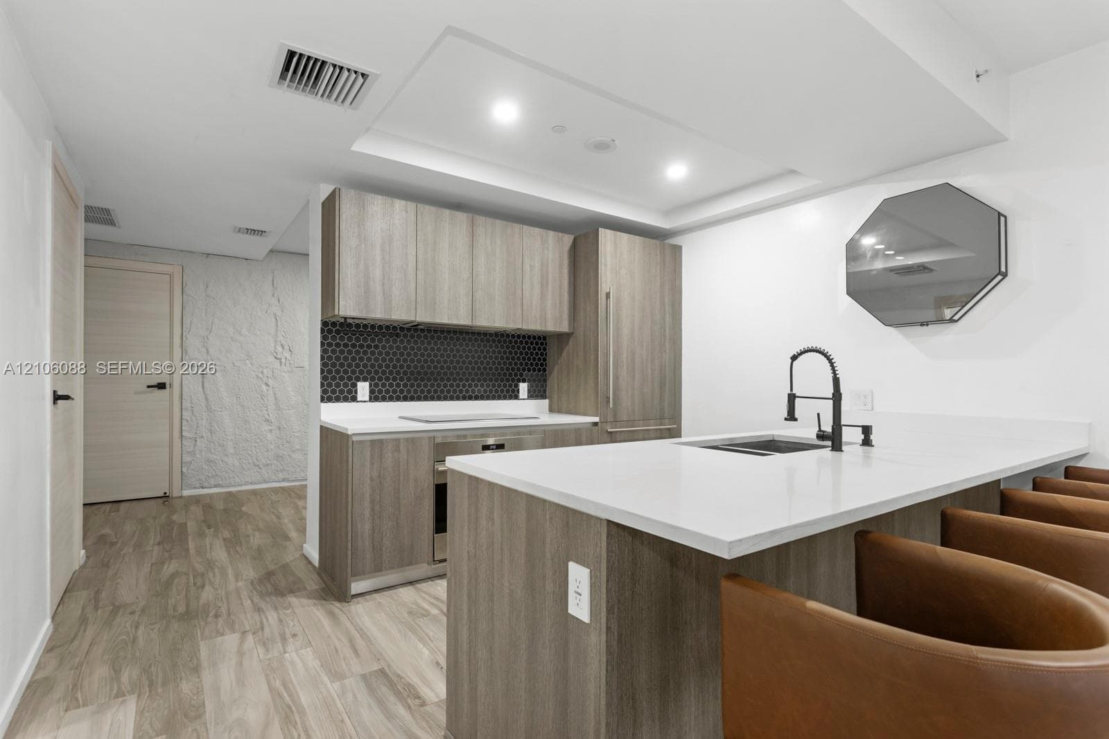 Contemporary kitchen featuring wood-finish cabinetry, white solid-surface countertops, a black hexagonal tile backsplash, a matte black kitchen faucet, and wood-finish flooring