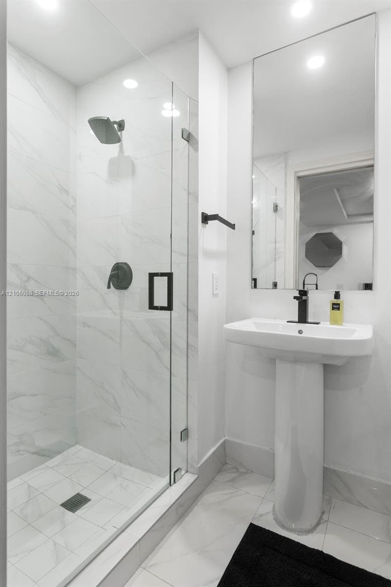 Walk-in shower with floor-to-ceiling veined tile, frameless glass enclosure, matte black fixtures, and a pedestal sink
