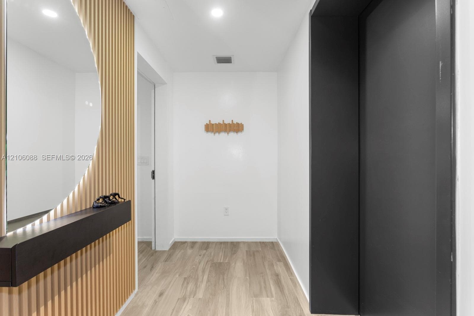 Foyer featuring wood-finish flooring, a vertical slat wood accent wall, a circular backlit wall mirror, and a sleek, matte black built-in floating shelf