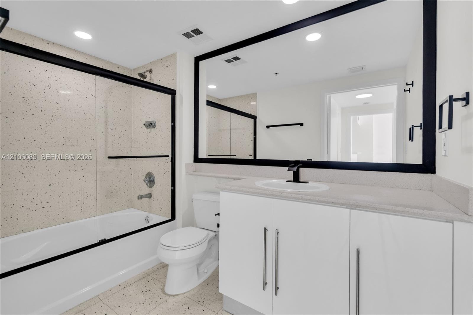 Bathroom featuring a white vanity cabinet with a stone countertop, an integrated sink with a matte black faucet, a large mirror with a black frame, and a bathtub with a glass enclosure and speckled tile surround