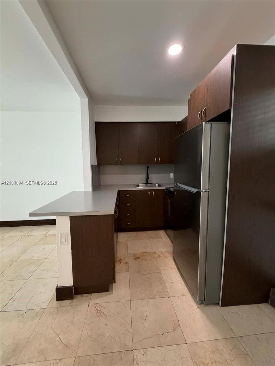 Kitchen featuring dark wood-finish cabinetry, solid-surface countertops, stone-finish flooring, and a stainless steel refrigerator