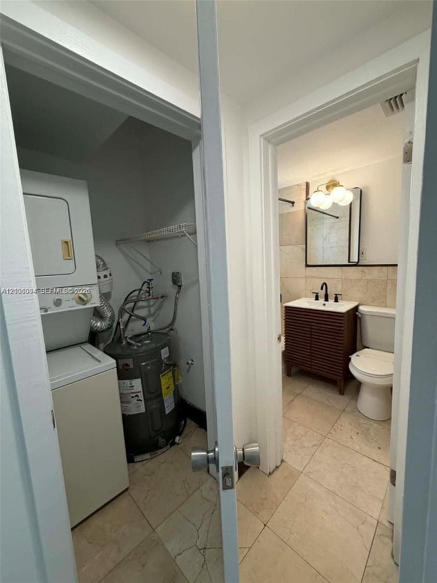 Laundry closet with stacked washer and dryer unit, wood-finish vanity with rectangular vessel sink, matte black faucet, wall-mounted globe lighting, and tile flooring