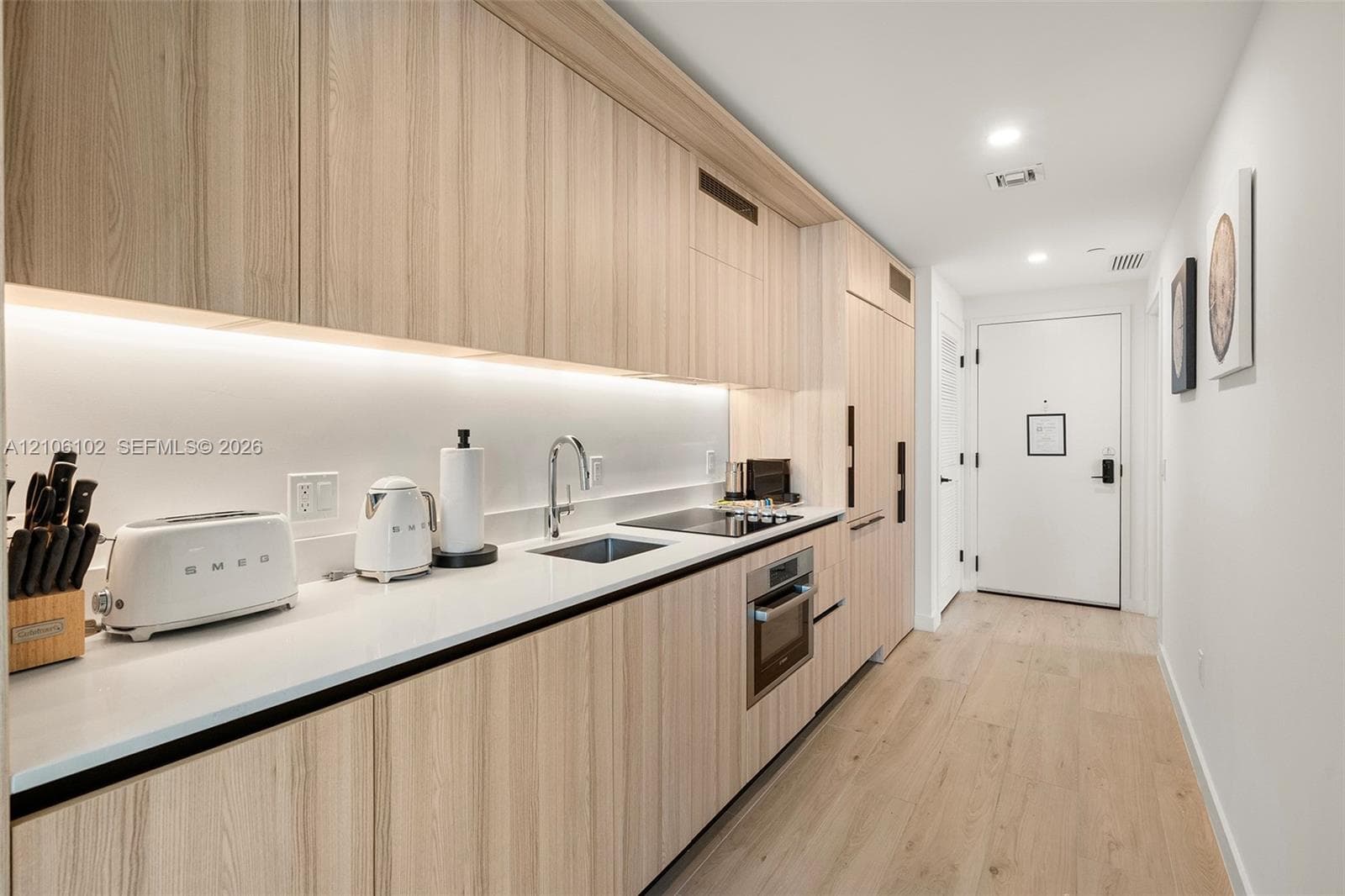Kitchen featuring light wood-finish cabinetry, solid surface countertops, integrated appliances, under-cabinet lighting, and wood-finish flooring