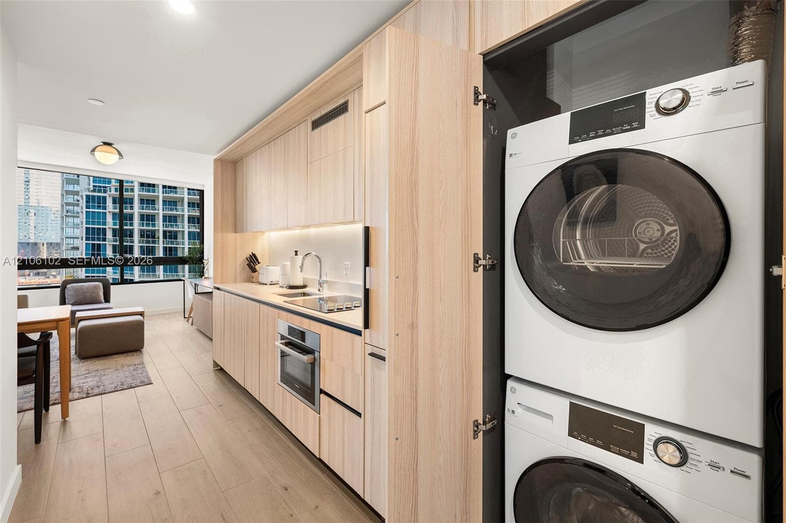 Kitchenette featuring light-toned cabinetry, built-in stacked laundry unit, integrated under-cabinet lighting, and wood-finish flooring