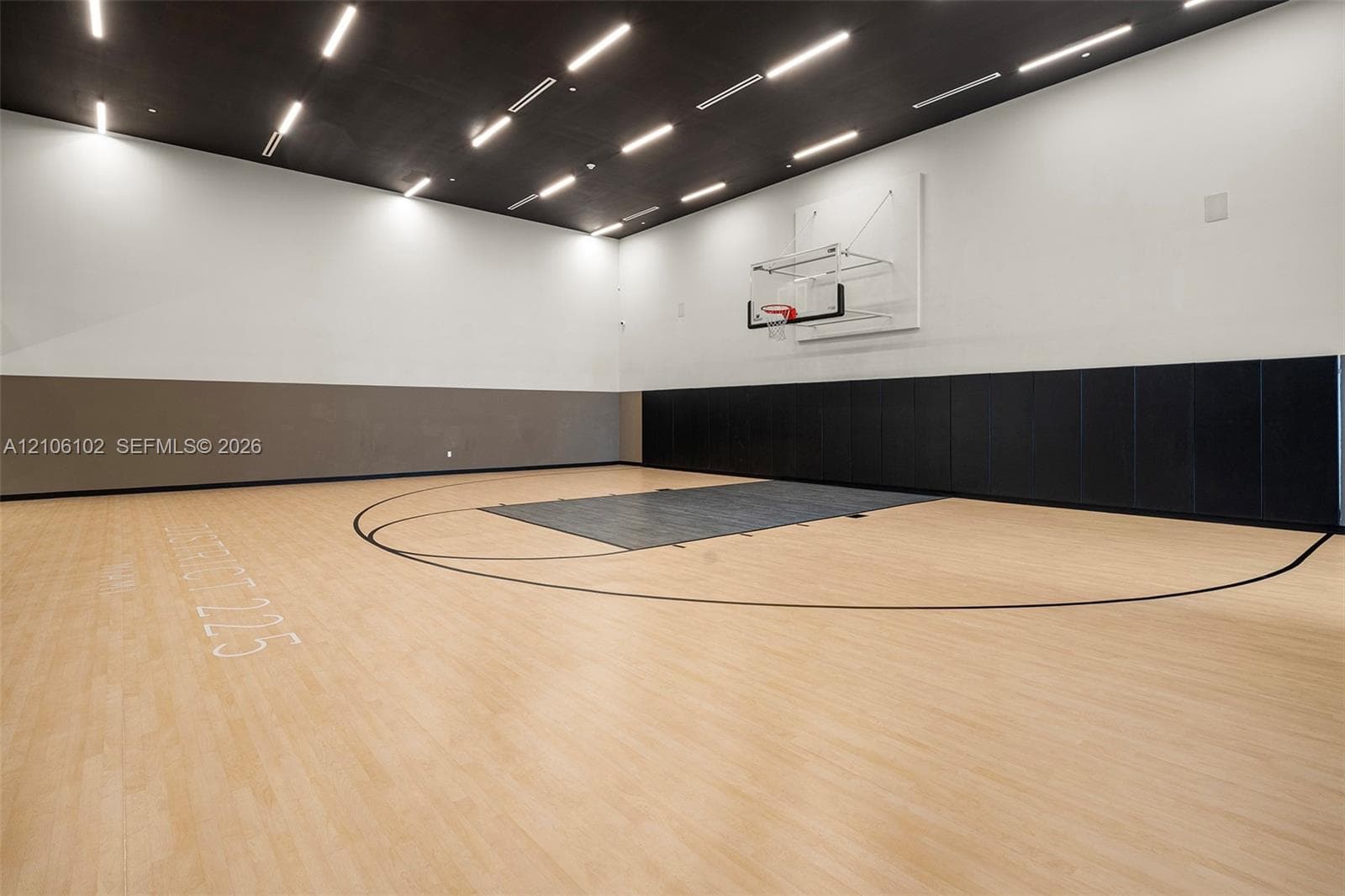 Indoor athletic court featuring wood-finish flooring, a wall-mounted basketball hoop, a black paneled ceiling with integrated linear lighting, and protective wall padding