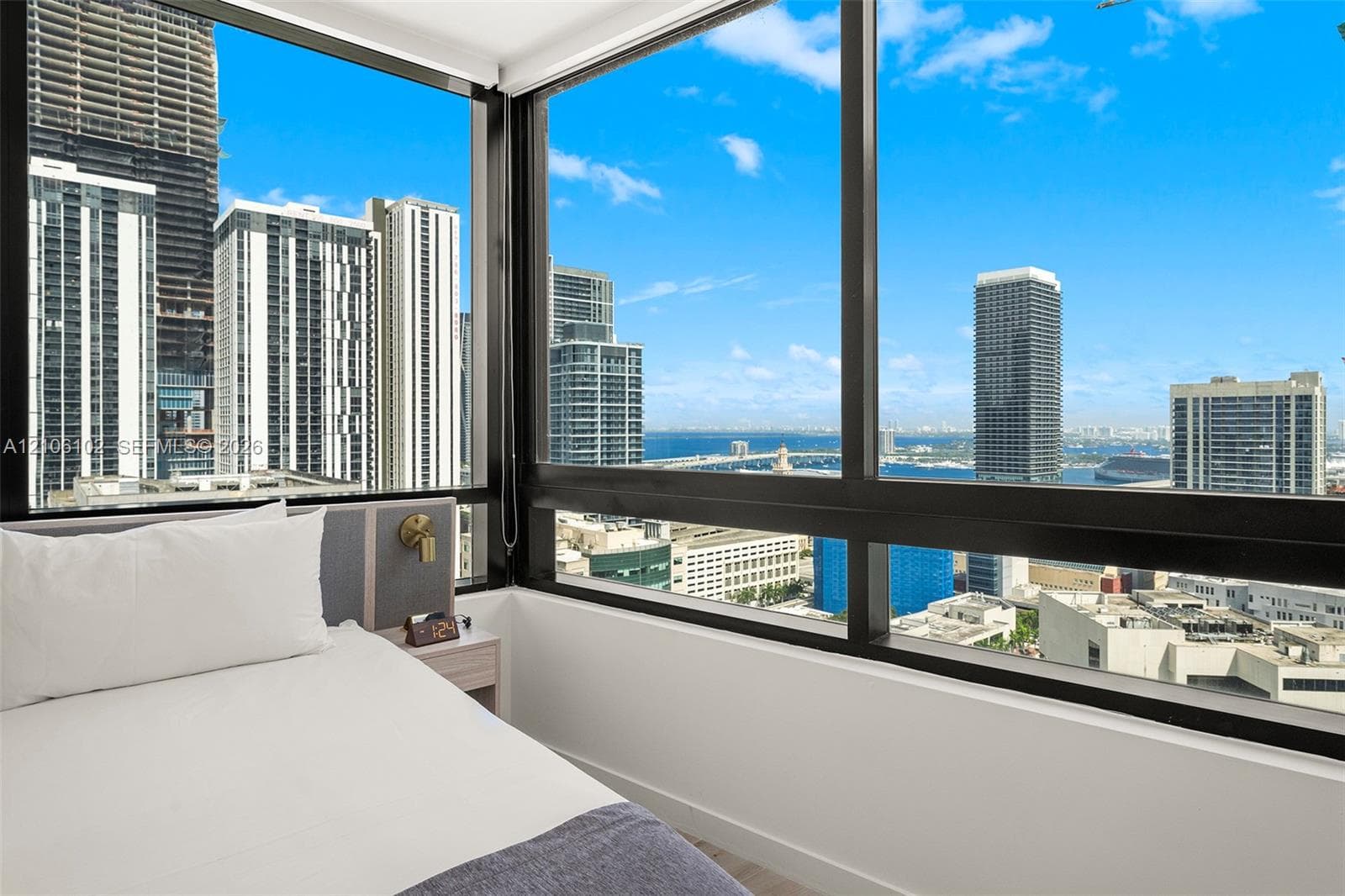 Floor-to-ceiling windows featuring panoramic urban and water scenery, black metal framing, and a built-in bedside panel with integrated lighting
