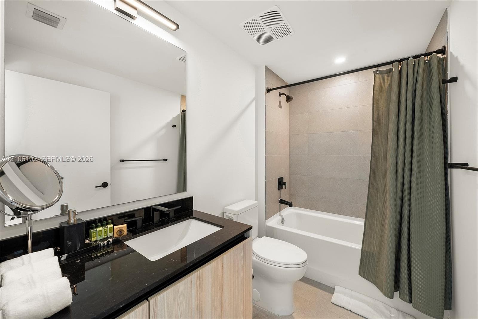 Vanity featuring a dark countertop, integrated rectangular sink, wood-finish cabinetry, and matte black fixtures, complemented by a tiled shower surround