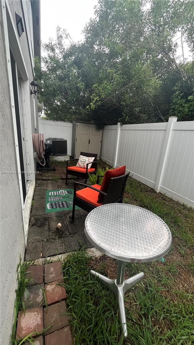 Outdoor area featuring a paved stone patio, white vinyl privacy fencing, and exterior wall-mounted lighting
