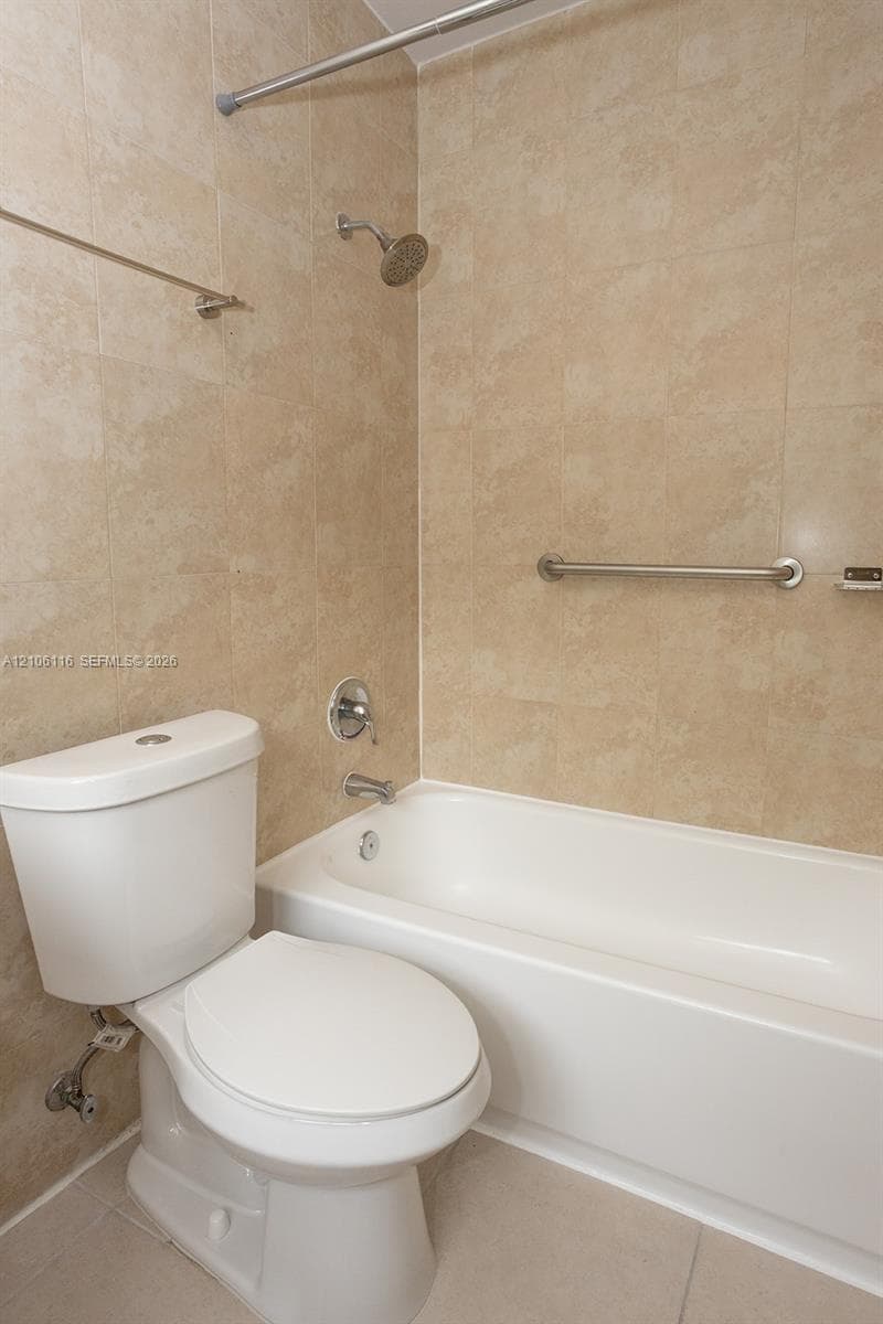 Bathroom featuring a white porcelain toilet, a white acrylic bathtub, neutral-toned tiled wall surrounds, a chrome shower head, and an installed chrome grab bar