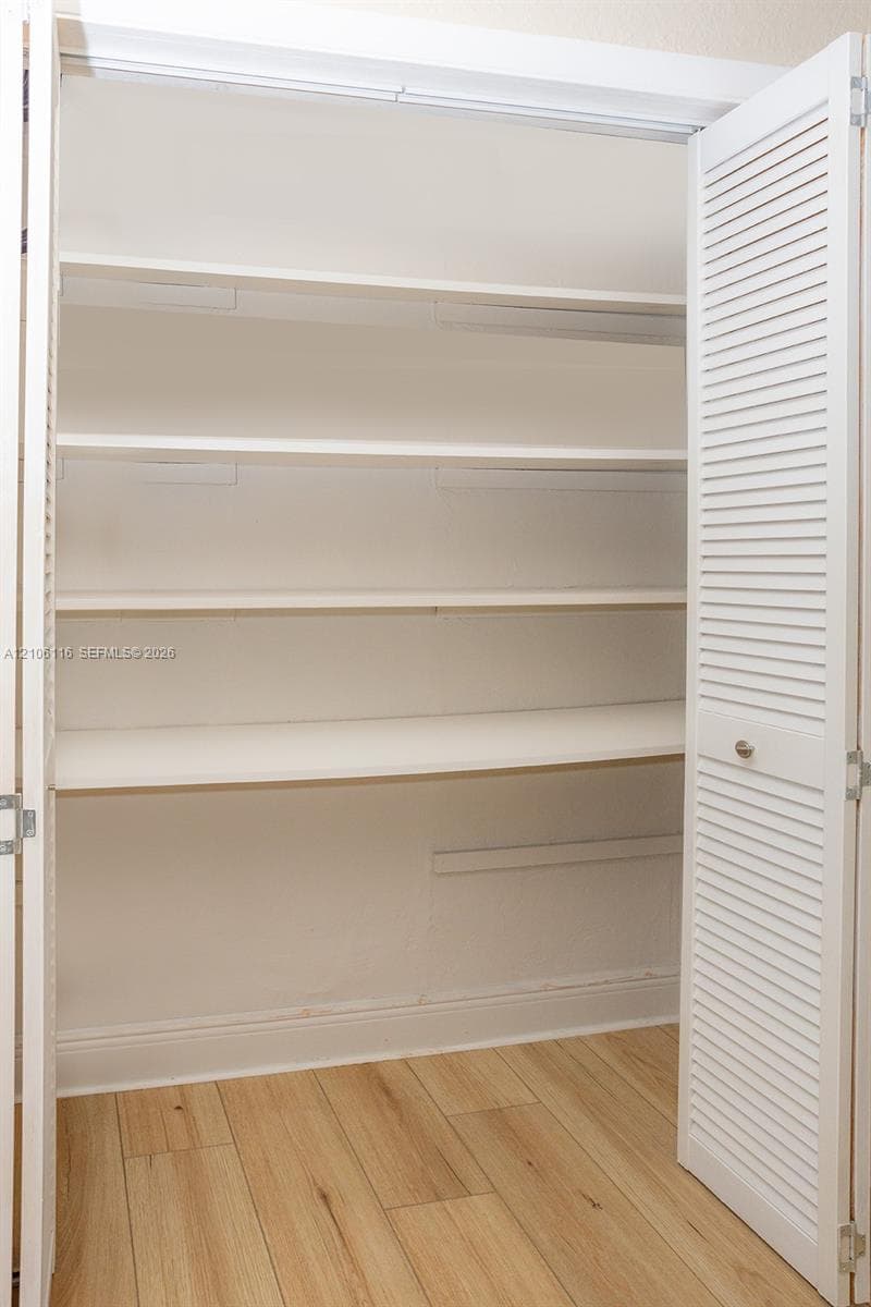 Built-in storage closet featuring white louvered doors and four interior shelves over wood-finish flooring