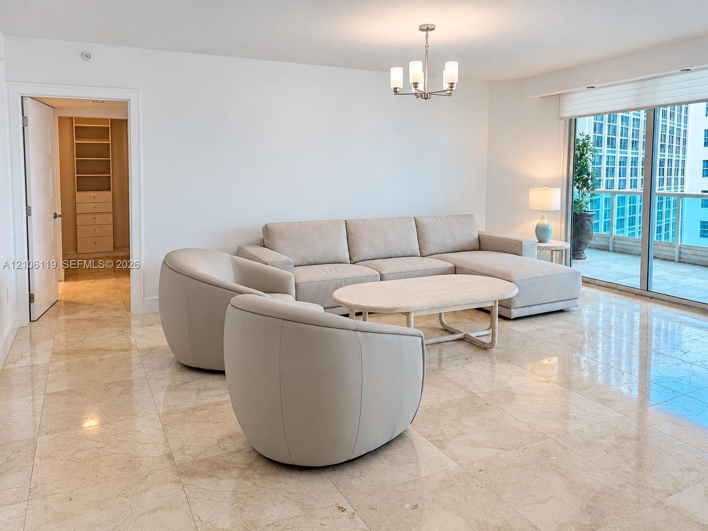 Open-concept interior featuring light-toned stone flooring, floor-to-ceiling glass sliding doors, a glass-railed balcony, and a multi-arm chandelier