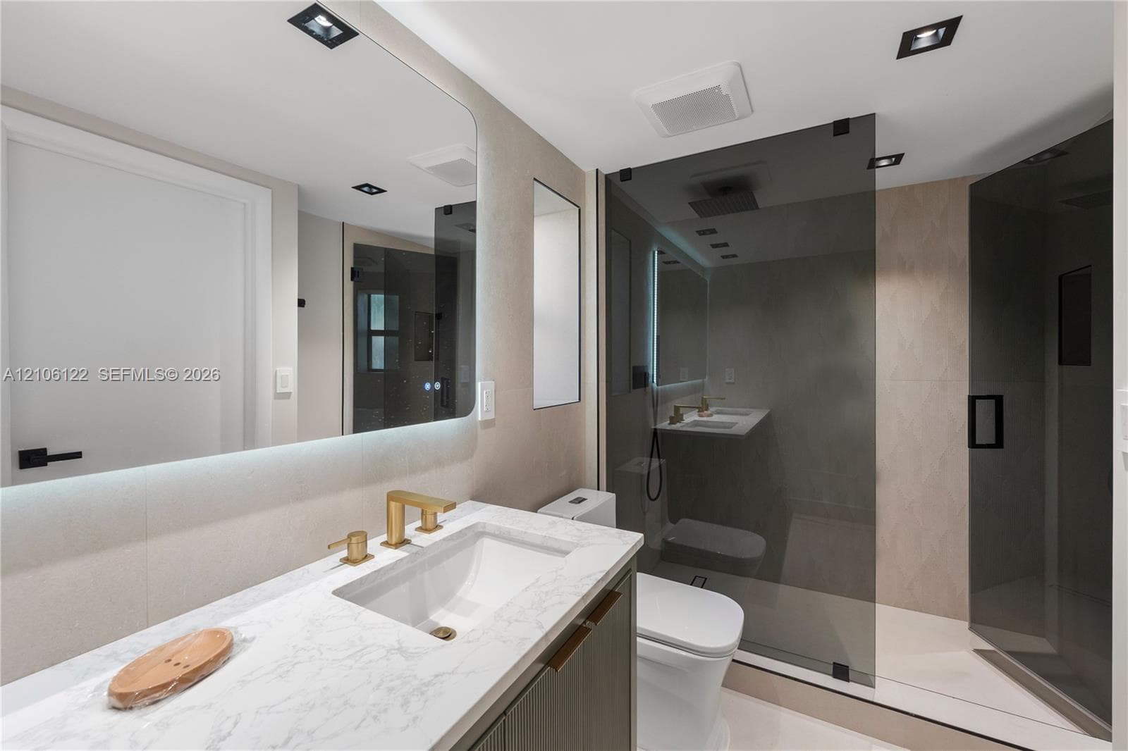 Bathroom featuring a floating vanity with a stone-finish countertop, undermount sink, brass-tone faucet, large LED-backlit mirror, and a walk-in shower with a glass enclosure and rainfall showerhead