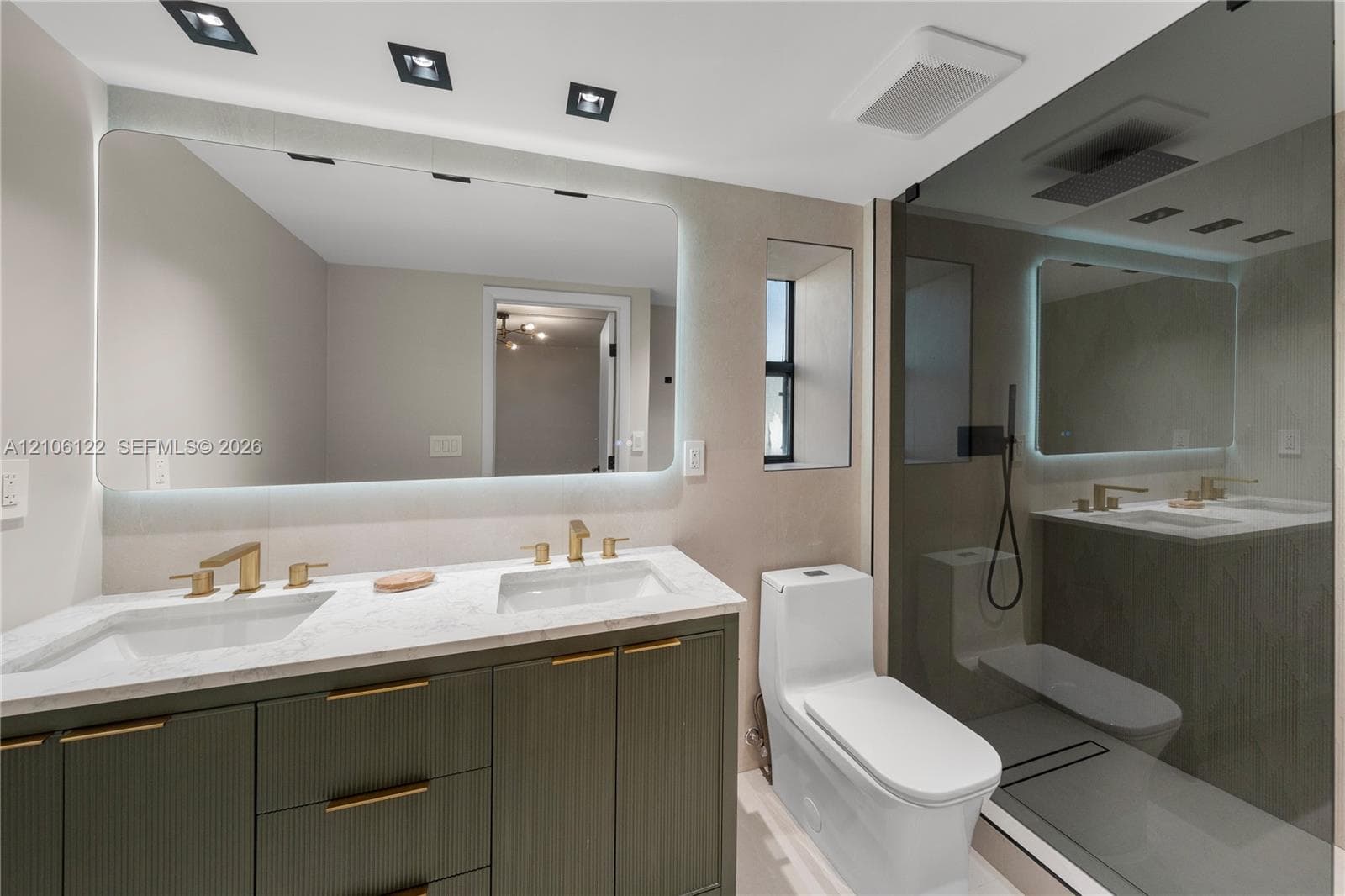 Dual-sink vanity with light-toned stone countertop, ribbed-texture cabinetry, integrated LED-backlit mirror, and brushed gold-tone fixtures, complemented by a glass-enclosed shower with rainfall showerhead and recessed ceiling lighting