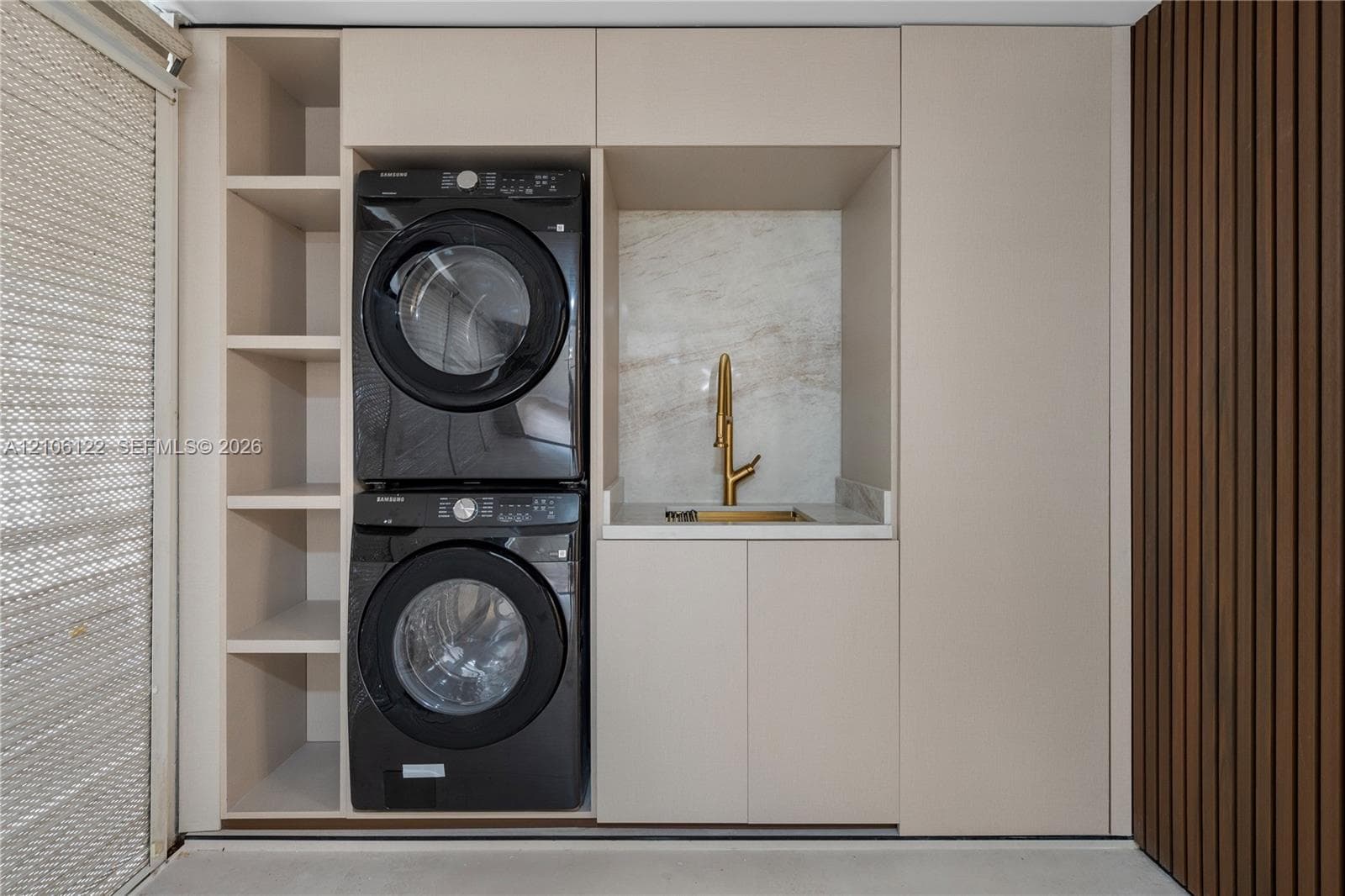 Integrated laundry area featuring custom cabinetry, stone-finish backsplash, gold-toned faucet, open shelving, and decorative wood-slat wall paneling