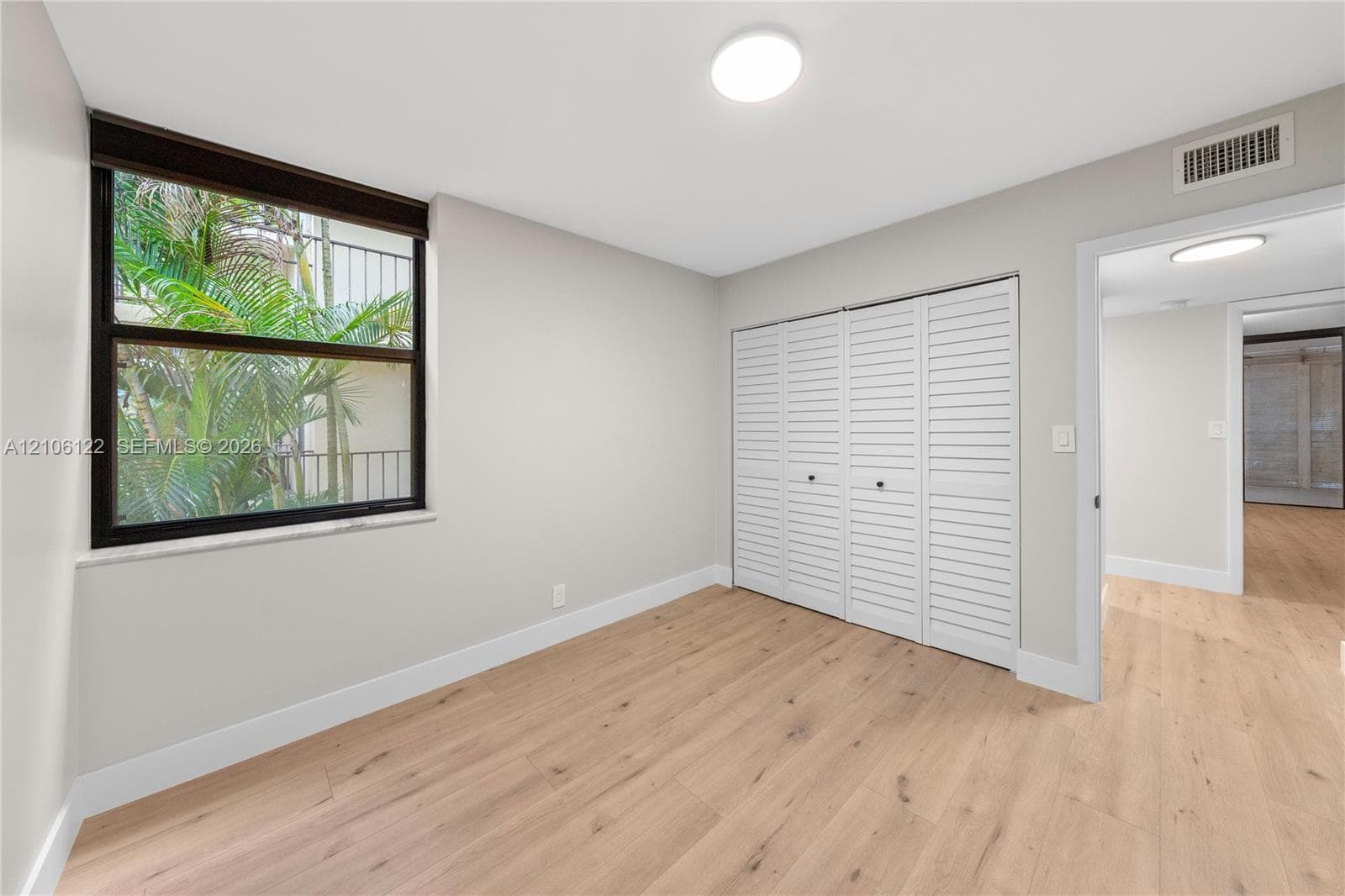 Room with wood-finish flooring, white louvered-style closet doors, neutral wall paint, and a window with dark-frame trim
