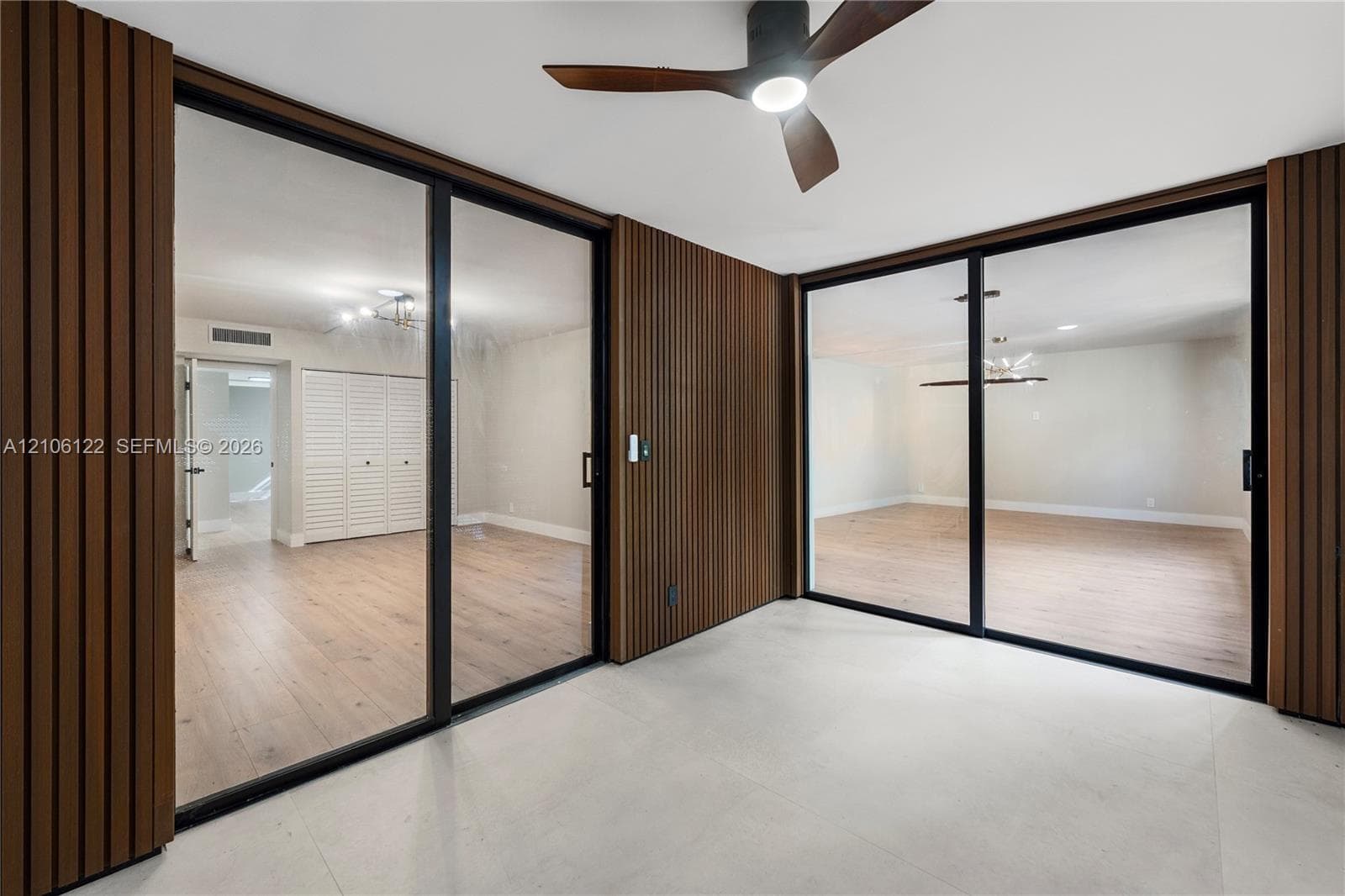 Slatted wood wall paneling, floor-to-ceiling glass sliding doors with black frames, wood-finish flooring, and a modern ceiling fan with integrated lighting