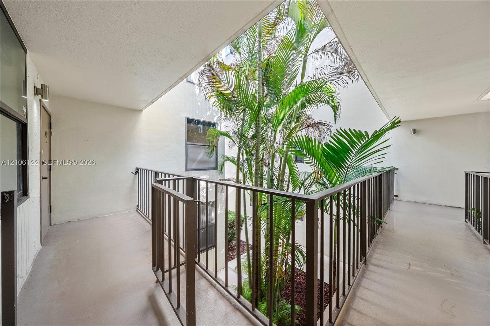 Open-air walkway featuring metal railings, concrete flooring, and a central atrium with mature palm trees
