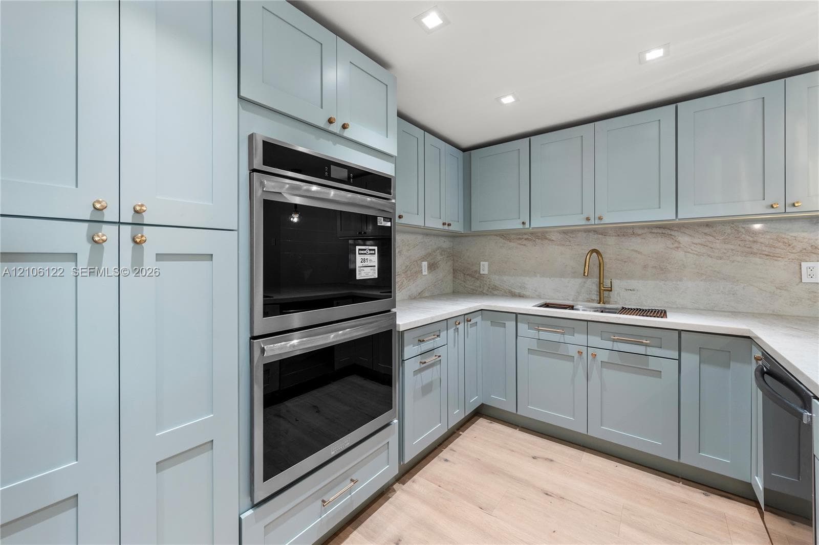Kitchen featuring custom soft-blue shaker cabinetry, stone backsplash, built-in double wall oven, gold-finish faucet, and wood-finish flooring