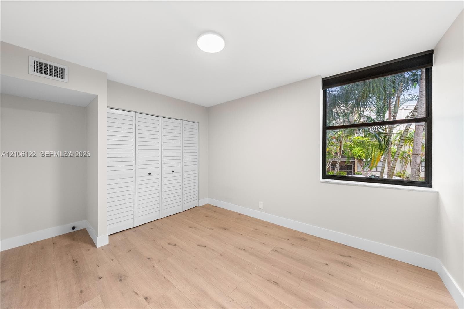 Neutral-toned room featuring wood-finish flooring, white louvered closet doors, and a large window with a black frame