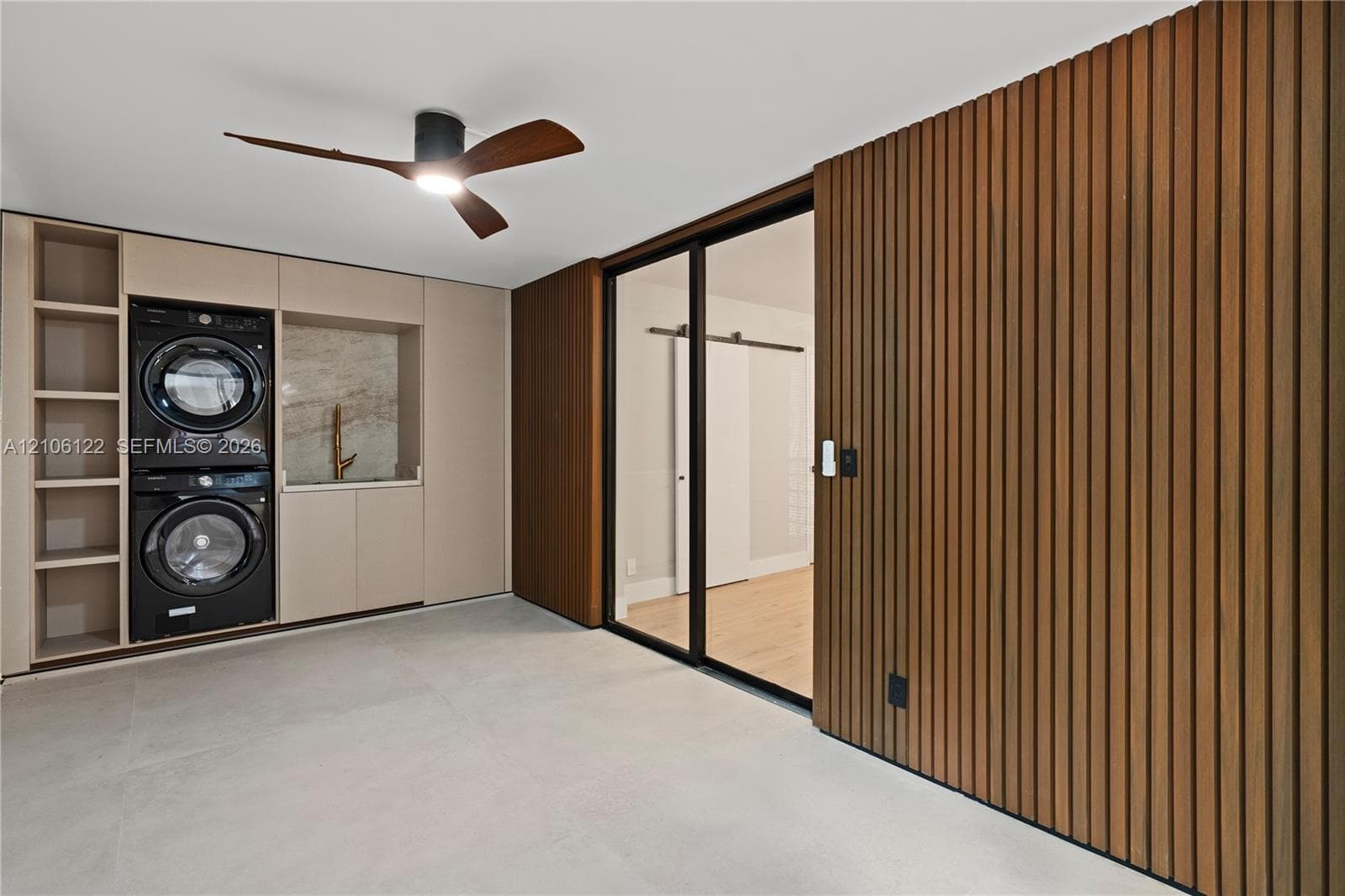 Built-in cabinetry with integrated appliance housing, vertical wood-slat wall paneling, full-height glass-paneled door with black frame, matte-finish flooring, and minimalist ceiling fan with integrated lighting