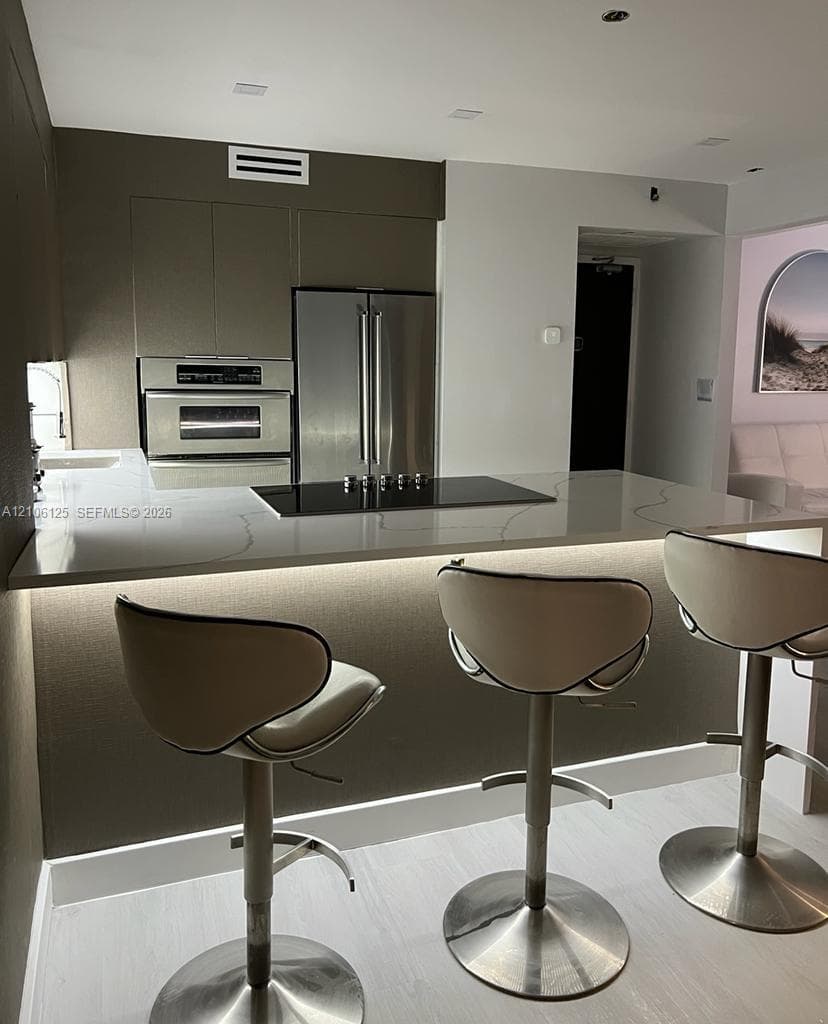 Kitchen featuring flat-panel cabinetry, stainless steel appliances including a wall-mounted oven, and a solid-surface waterfall island with an integrated cooktop and under-counter lighting