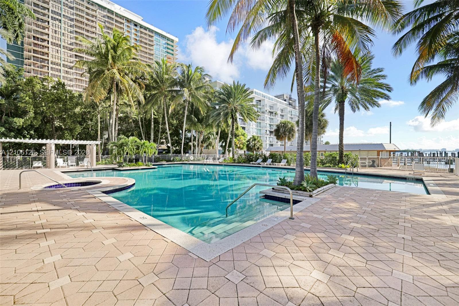 In-ground swimming pool featuring a tiled perimeter, integrated metal handrails, a covered pavilion, and surrounding paver decking lined with mature palm trees