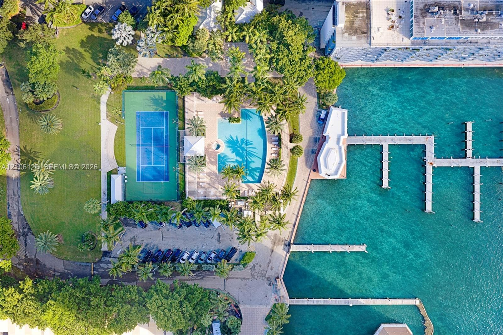 Aerial view of recreational amenities featuring a paved swimming pool deck, integrated spa, hard-surface tennis court, and mature palm landscaping