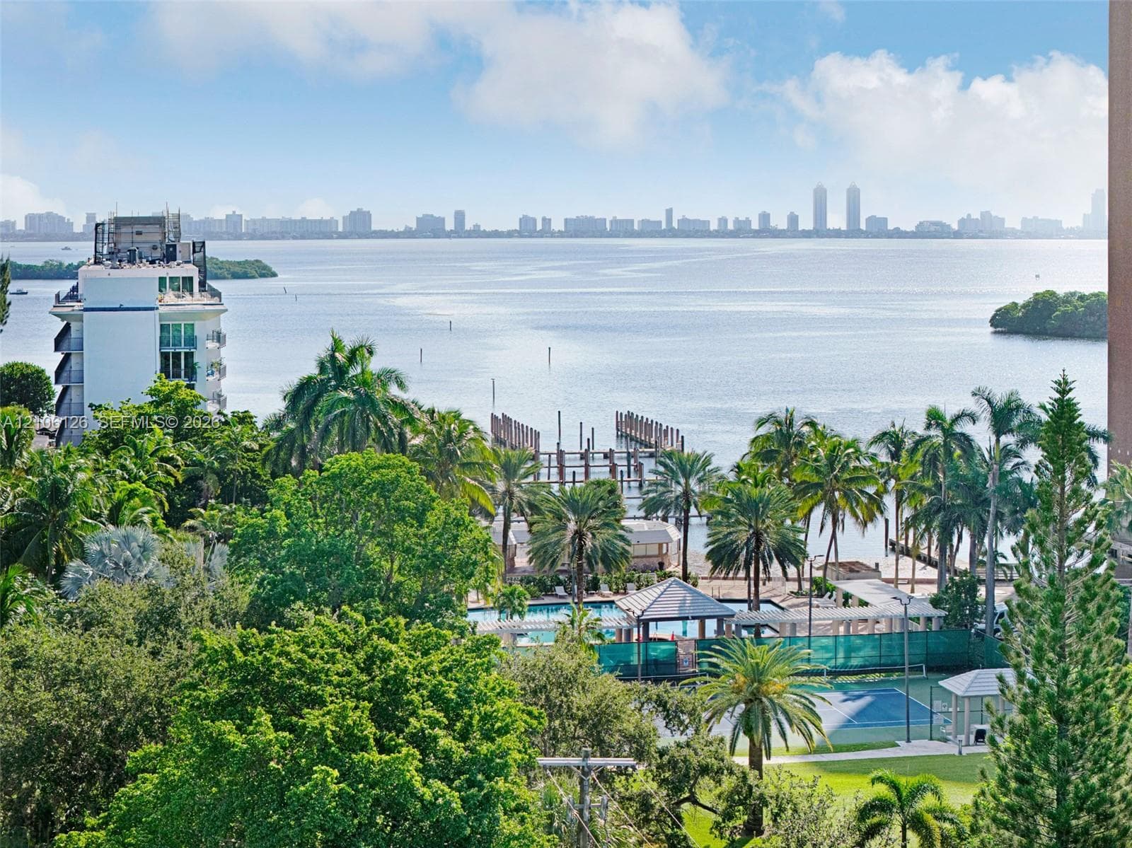 Panoramic water and skyline backdrop featuring mature tropical landscaping, an outdoor swimming pool, and a tennis court