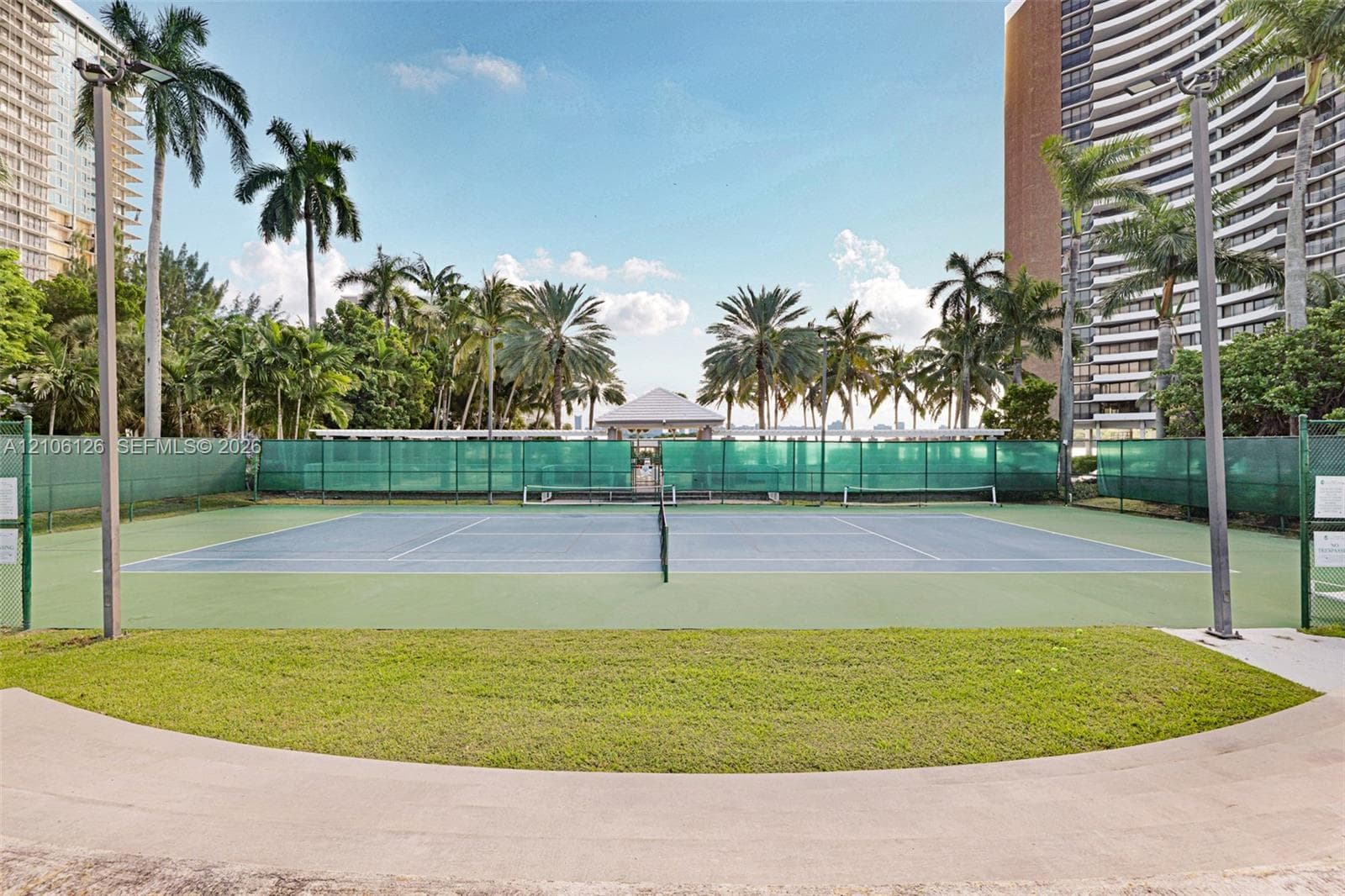 Hard-surfaced tennis court featuring a blue and green color scheme, perimeter fencing, and surrounding mature palm trees