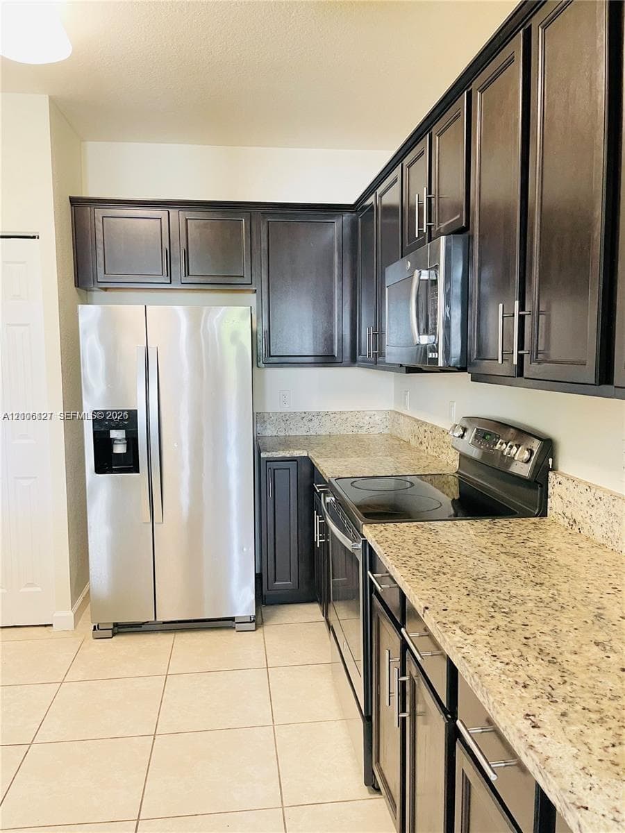 Kitchen featuring dark wood cabinetry, speckled stone countertops, tile flooring, and stainless steel appliances including a built-in microwave and refrigerator