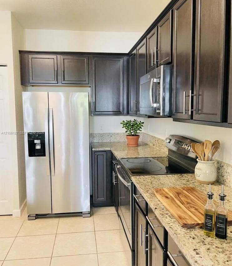 Kitchen featuring dark-finish cabinetry, stone-surface countertops, tile flooring, and stainless steel appliances including a built-in microwave