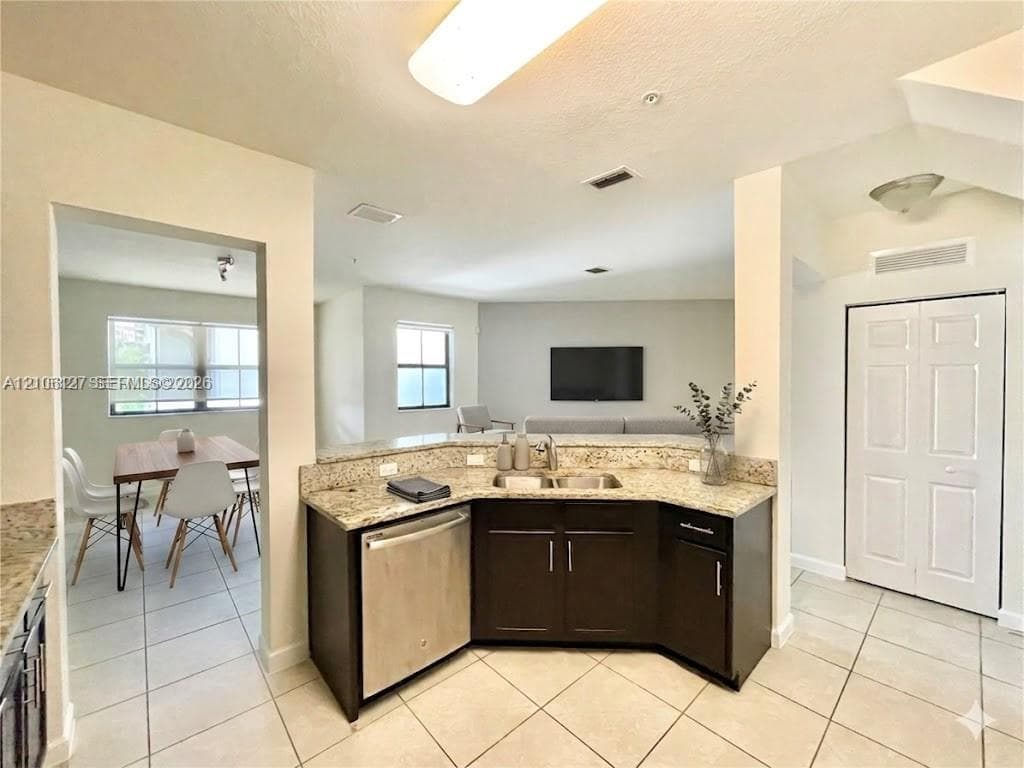 Kitchen featuring dark cabinetry, stone-finish countertops, an undermount double-basin sink, and stainless steel dishwasher, complemented by light-toned tile flooring