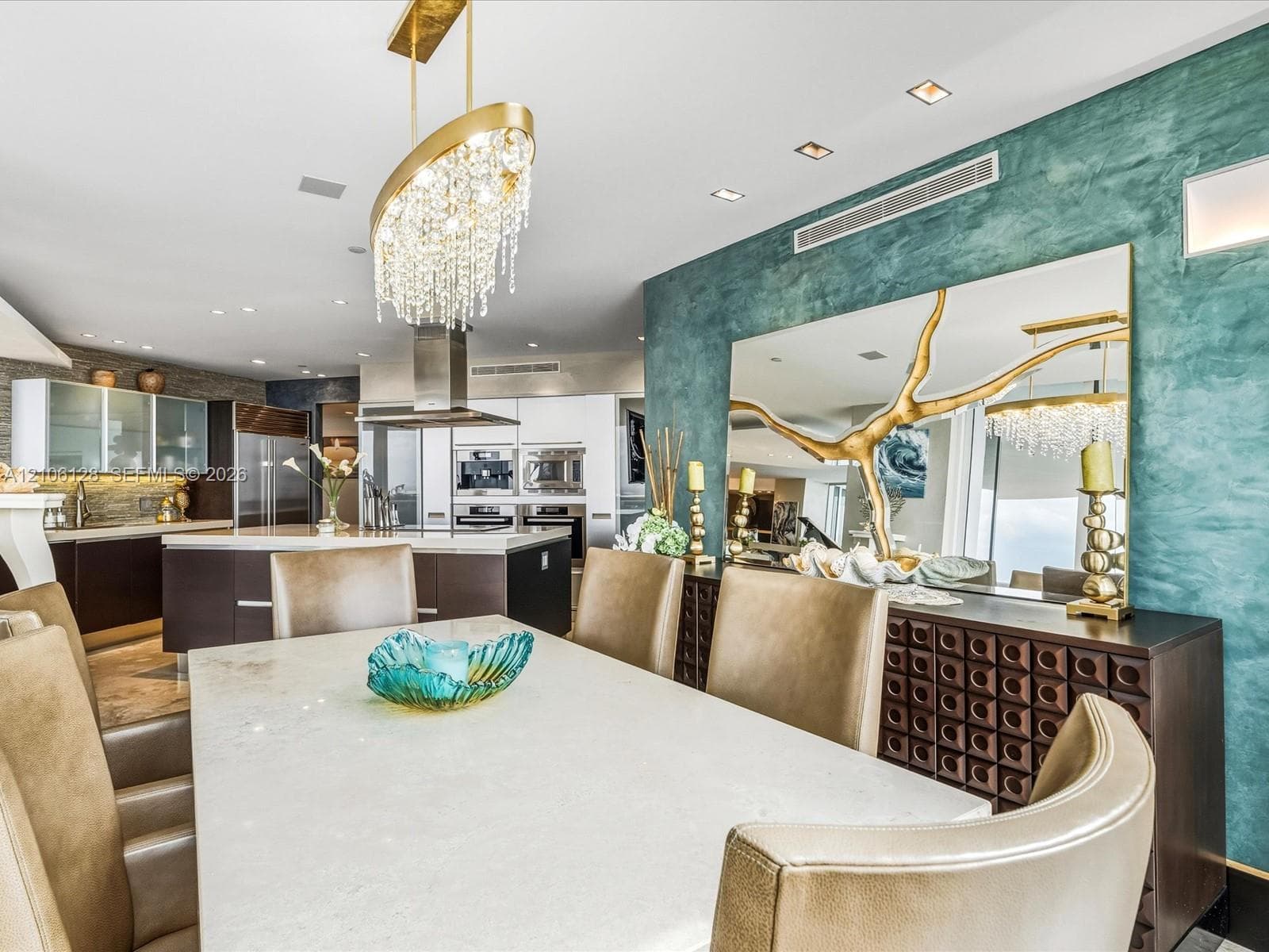 Dining area featuring a custom crystal chandelier, textured teal feature wall, integrated recessed lighting, and a neutral stone-surface table