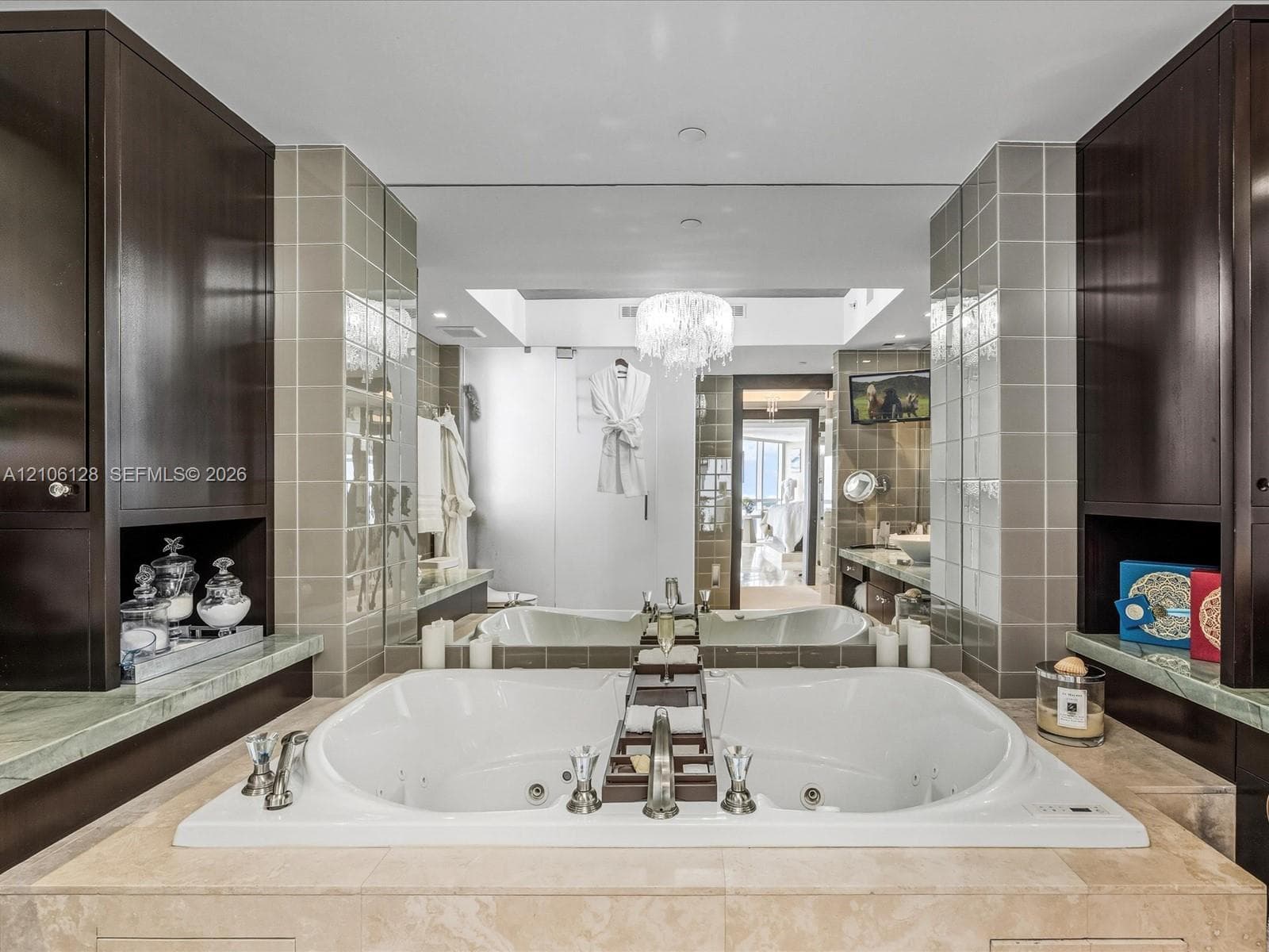 Jetted soaking tub with deck-mounted chrome fixtures, flanked by dark wood cabinetry, integrated stone countertops, and full-wall mirror accented by tile surround and a central crystal-style chandelier