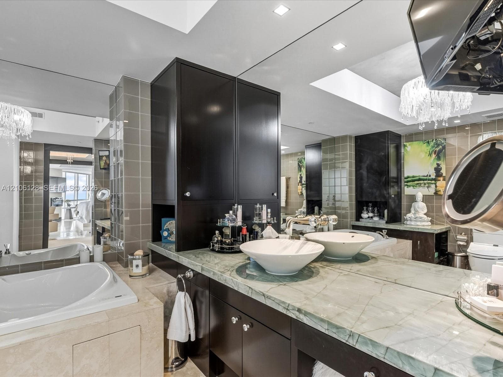 Custom dual-sink vanity featuring stone-surfaced countertops, dark built-in cabinetry, white vessel sinks, and a drop-in soaking tub encased in stone tile
