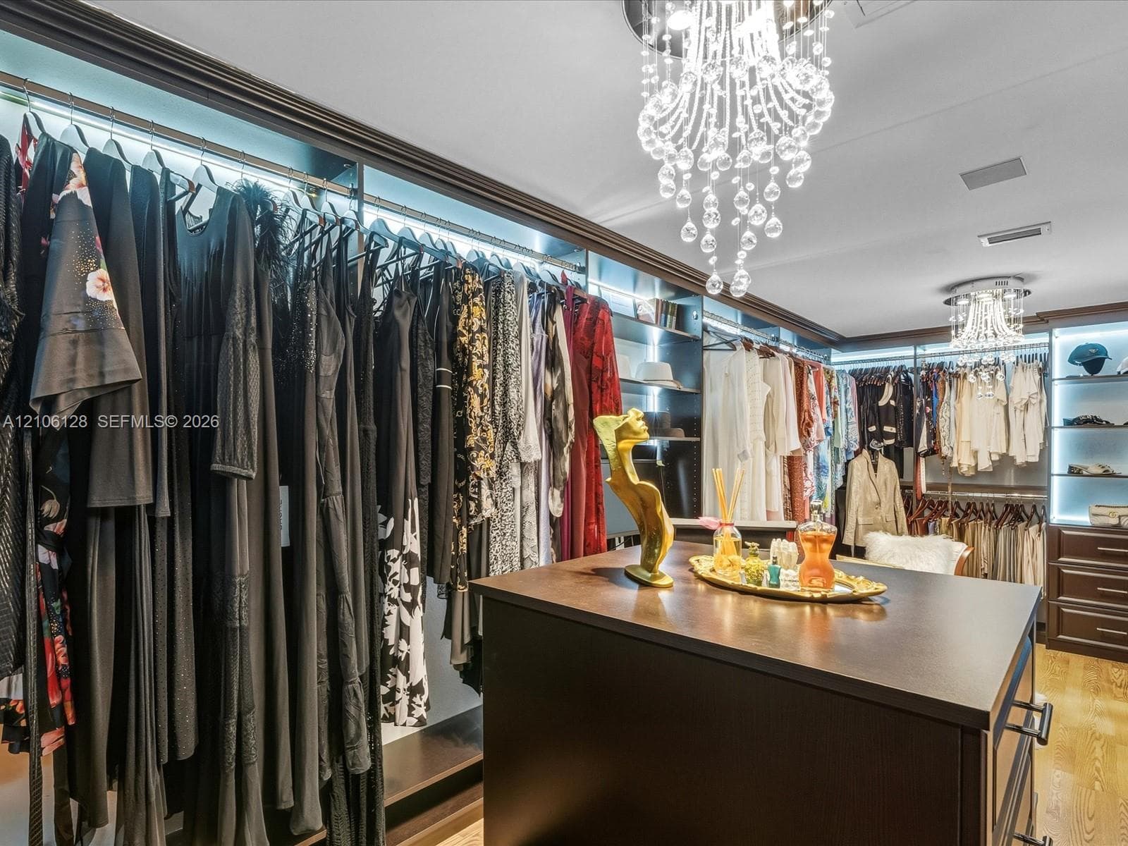 Custom walk-in closet featuring extensive dark-finish built-in shelving, integrated cabinet lighting, a central storage island, and two tiered crystal chandeliers