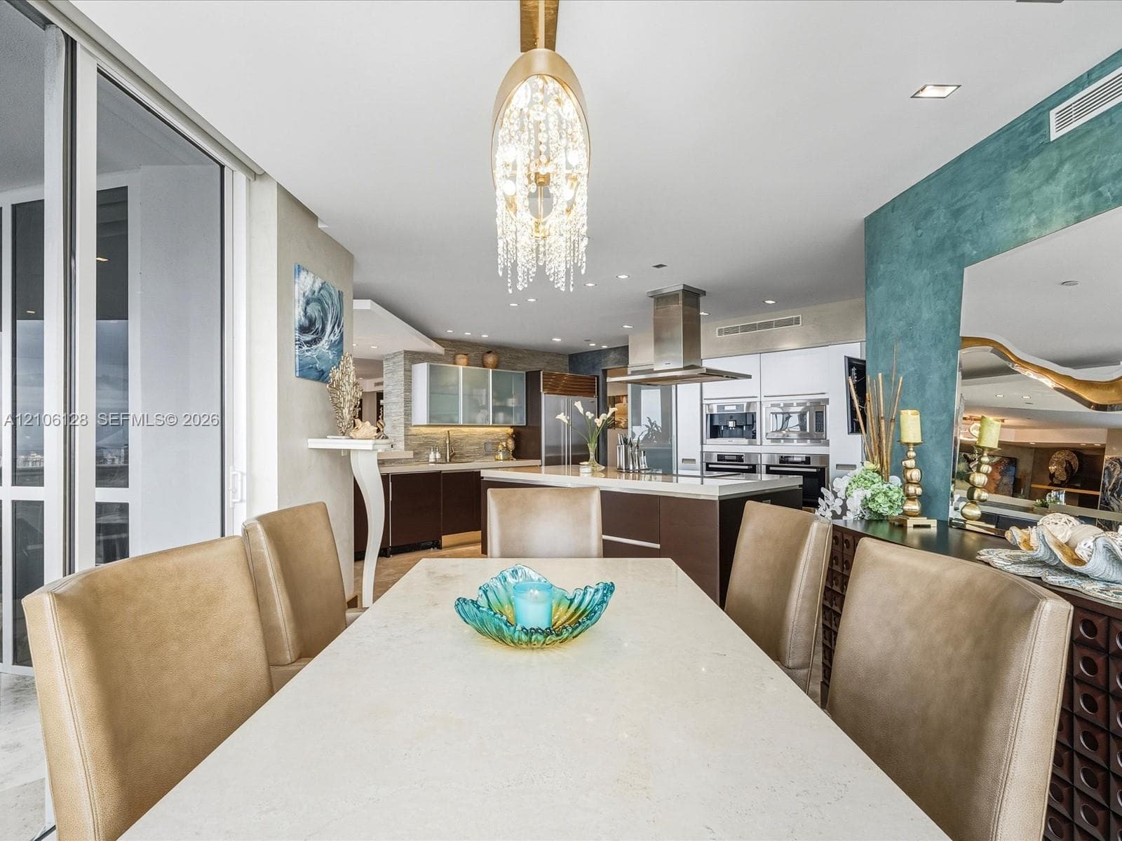 Open-concept interior featuring a stone-topped dining area, a tiered crystal chandelier, custom flat-panel cabinetry, a stainless steel chimney hood, and floor-to-ceiling glass paneling