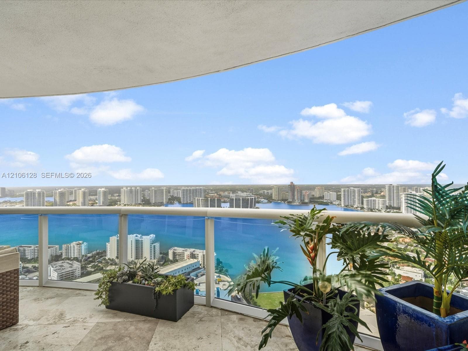 Curved balcony featuring natural stone flooring and a glass railing system, overlooking expansive water and coastal city scenery