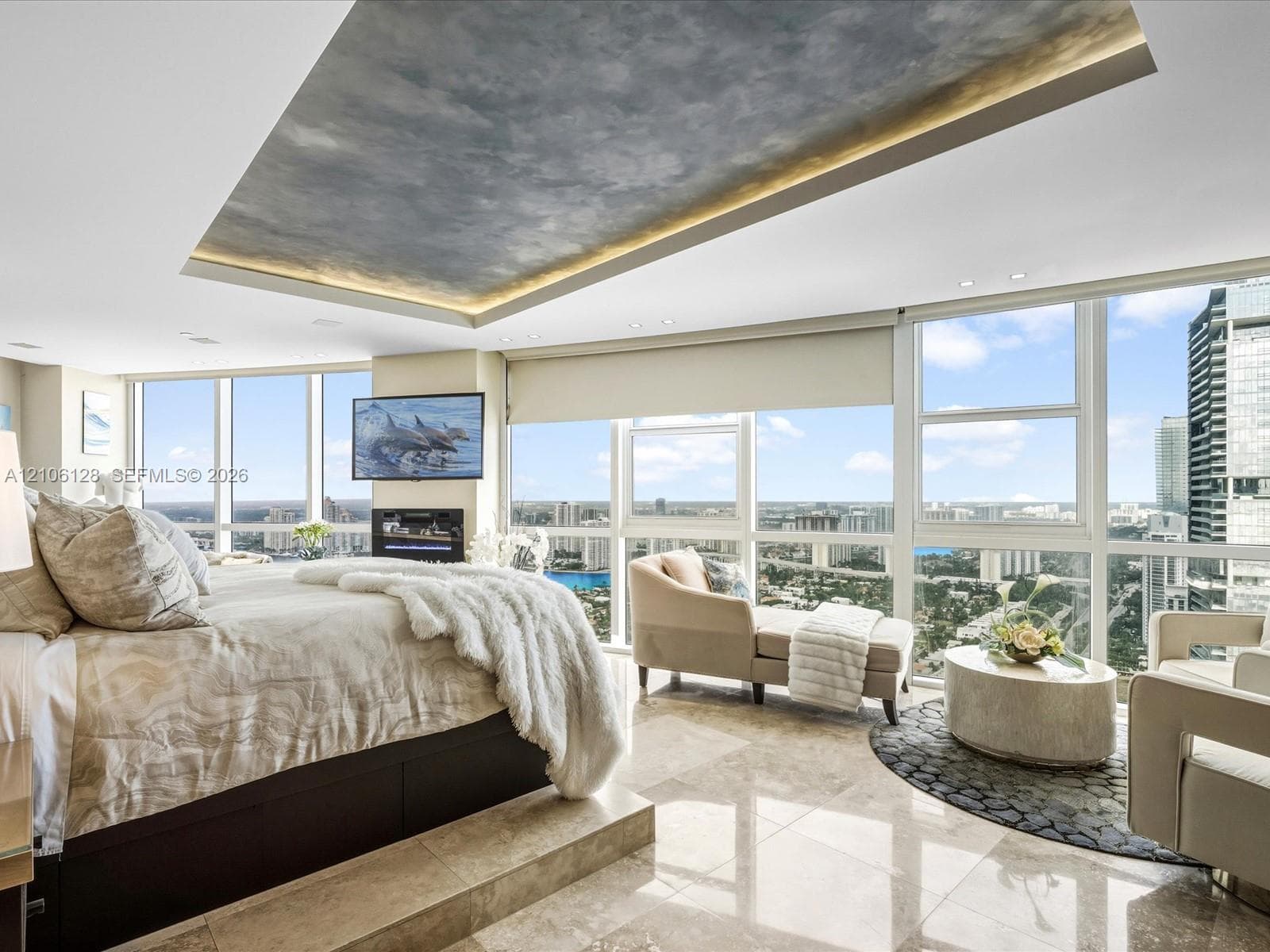 Floor-to-ceiling windows, polished stone flooring, recessed tray ceiling with integrated cove lighting, and a wall-mounted electric fireplace