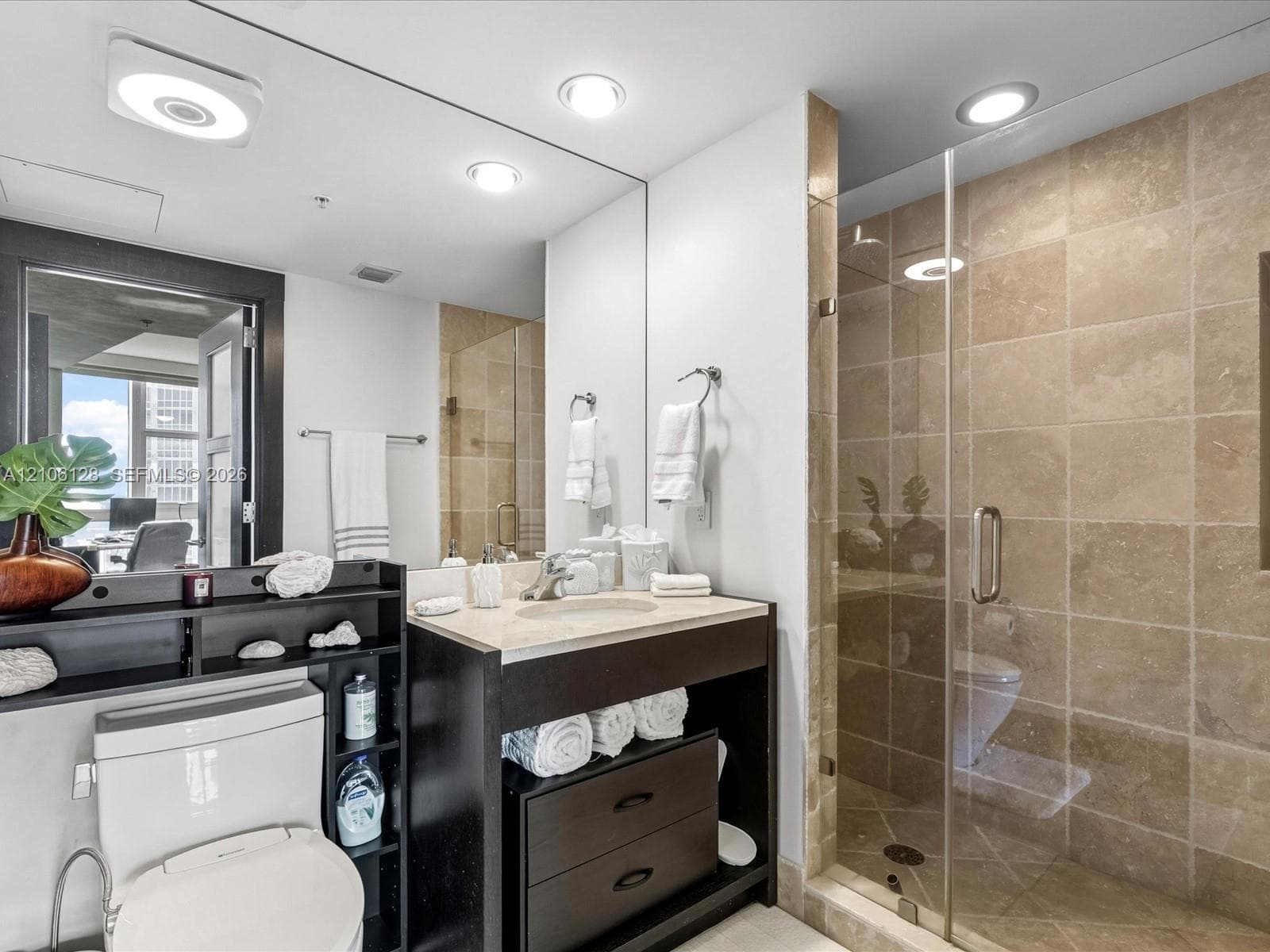 Bathroom featuring a vanity with stone countertop, under-mount sink, dark wood-finish cabinetry, full-width mirror, and a walk-in shower with tile surround and glass enclosure