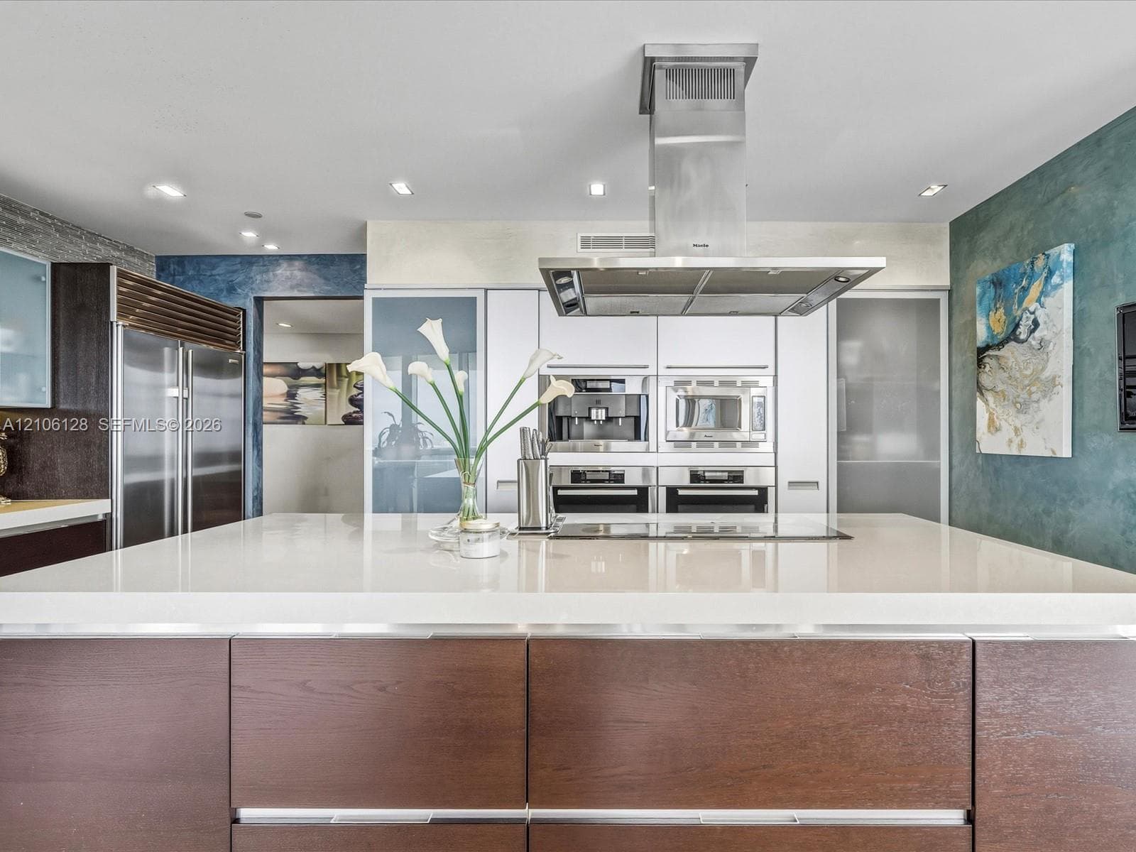 Kitchen featuring a large central island with a quartz countertop, flat-panel wood-finish cabinetry, built-in stainless steel appliances, and a ceiling-mounted stainless steel range hood