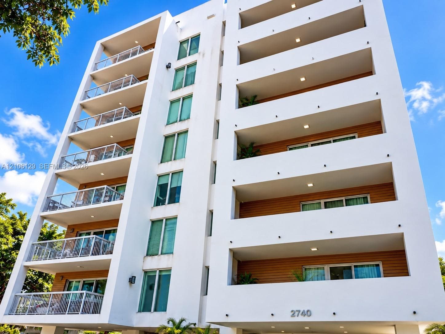 Mid-rise residential building featuring a smooth stucco facade, multi-story private balconies with glass railings, wood-finish exterior siding, and recessed ceiling lighting