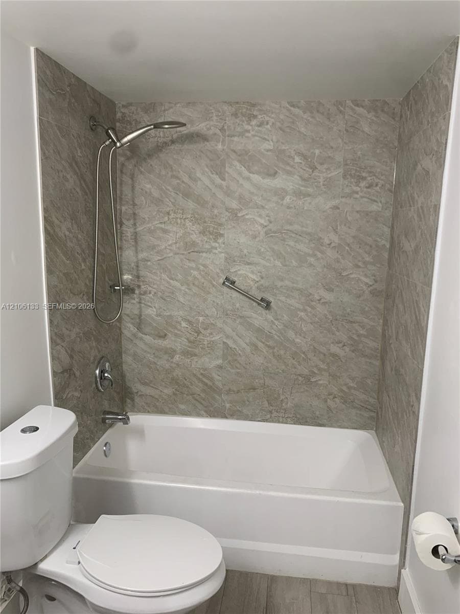 Full-size bathtub with stone-pattern tile surround, brushed nickel adjustable handheld shower fixture, built-in grab bar, and dual-flush toilet