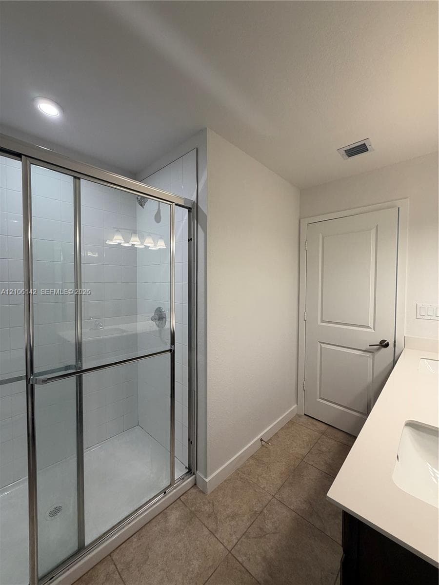 Walk-in shower with chrome-framed glass enclosure and tiled surround, dual-sink vanity with solid-surface countertop, and ceramic tile flooring