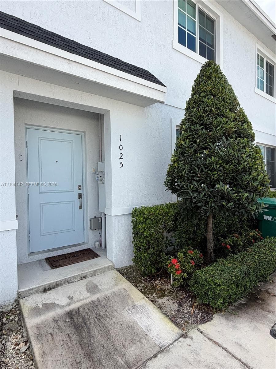 White stucco exterior featuring a recessed entryway, light blue paneled door, concrete walkway, and landscaped shrubbery