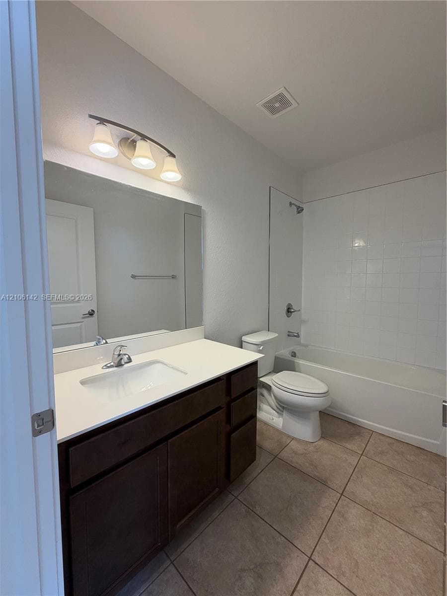Dark wood-finish cabinetry, white countertop with integrated sink, large wall-mounted mirror, three-light vanity fixture, and tiled tub-shower surround